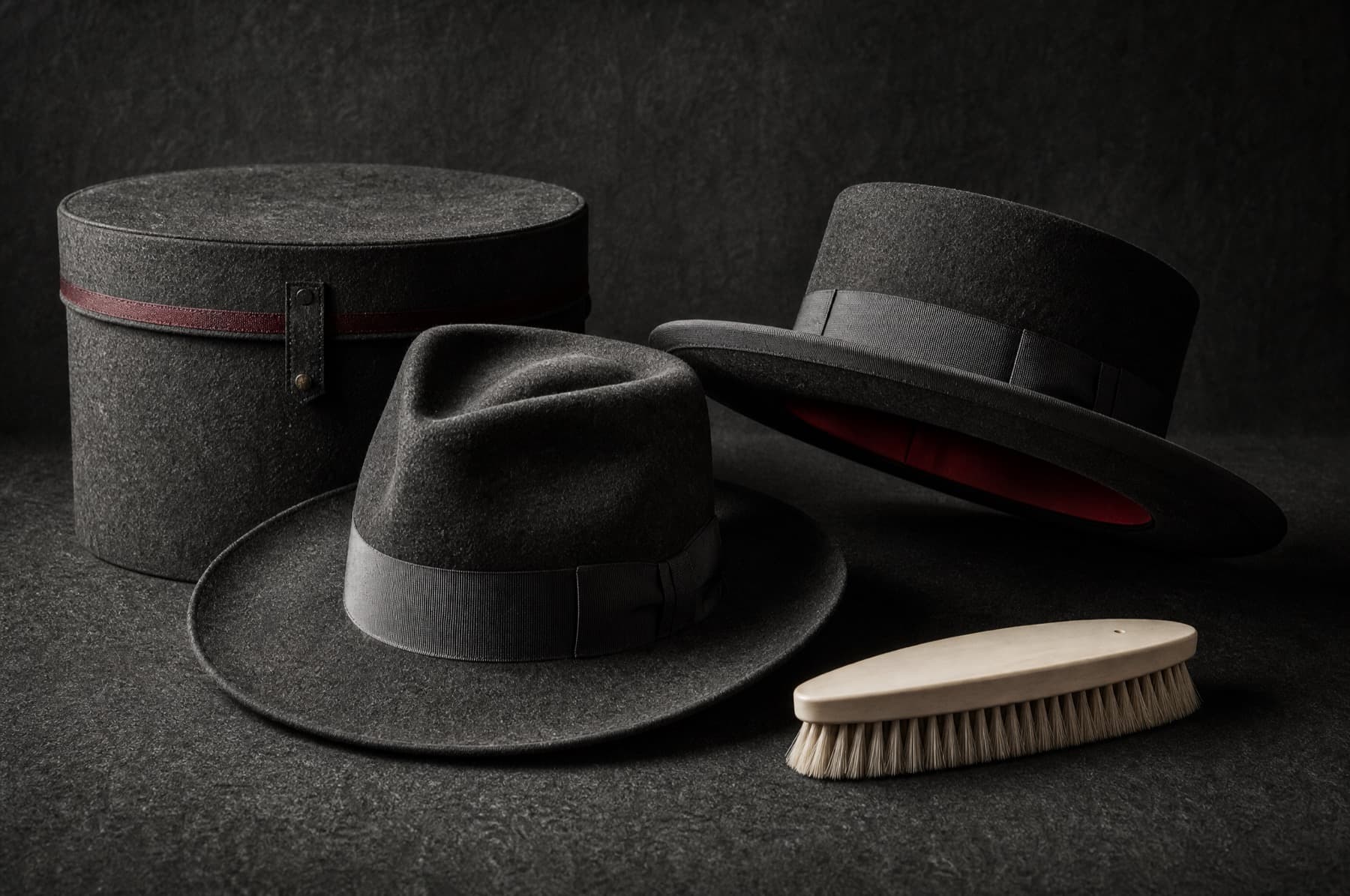 A soft-creased fedora and a rigid structured hat standing beside a hat box and brush